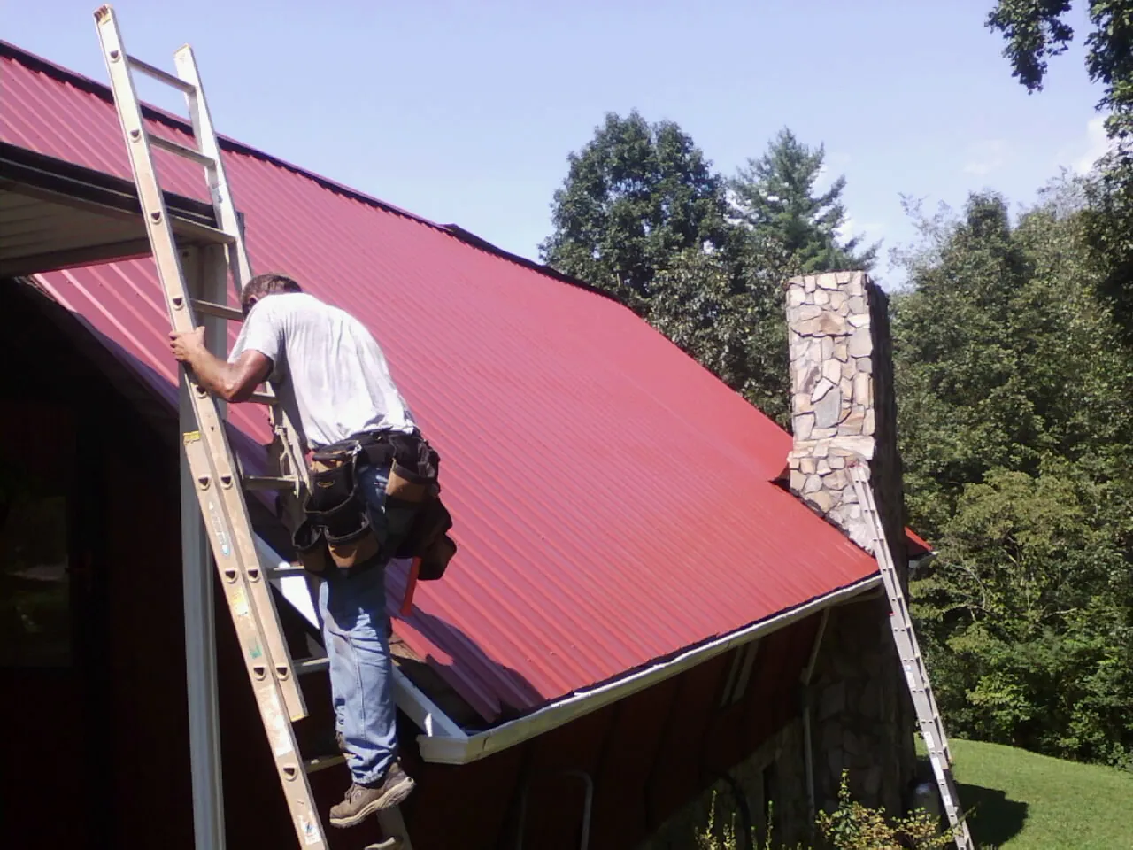Roofer on a ladder beside a red metal roof and stone chimney — Soffit & Fascia Repair by Roofing Brothers in Zolfo Springs